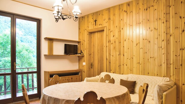 Una habitación acogedora con una mesa redonda, sillas, un sofá, un televisor en una estantería y una pared de madera con una cruz.