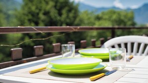 Una mesa de comedor con platos verdes, cubiertos y vasos, ubicada al aire libre con vistas a árboles y montañas.