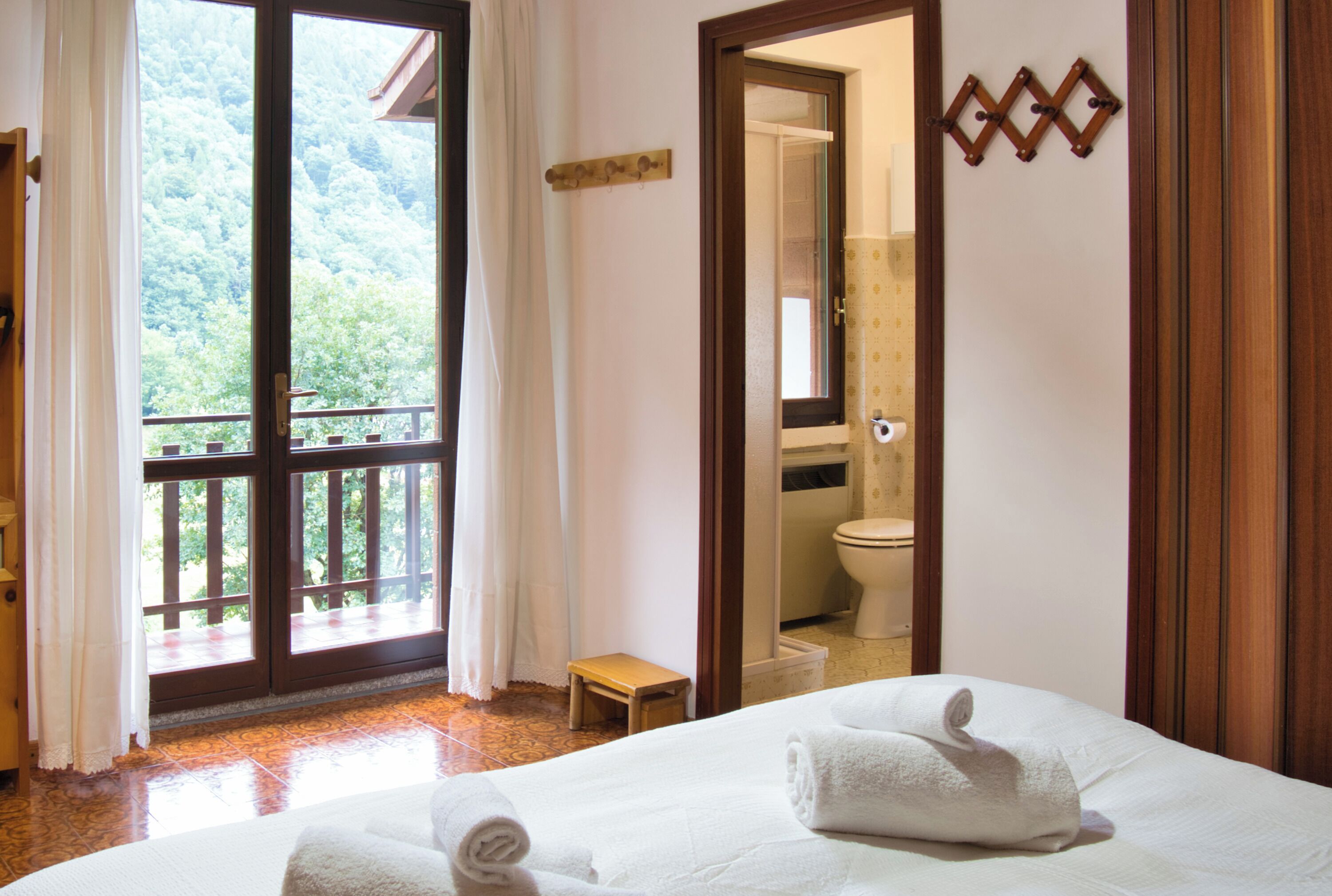 Een hotelkamer met een grote glazen schuifdeur die uitkomt op een balkon, een houten bankje, een bed met een opgerolde handdoek, en een badkamer zichtbaar in een spiegel.
