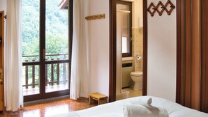 Habitación de hotel con un amplio ventanal corredero que da a un balcón, una banca de madera, una cama con una toalla enrollada y un baño visible a través de un espejo.