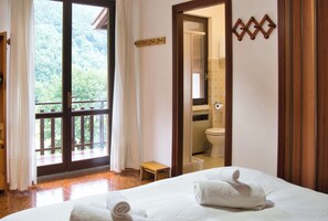 Een hotelkamer met een grote glazen schuifdeur die uitkomt op een balkon, een houten bankje, een bed met een opgerolde handdoek, en een badkamer zichtbaar in een spiegel.