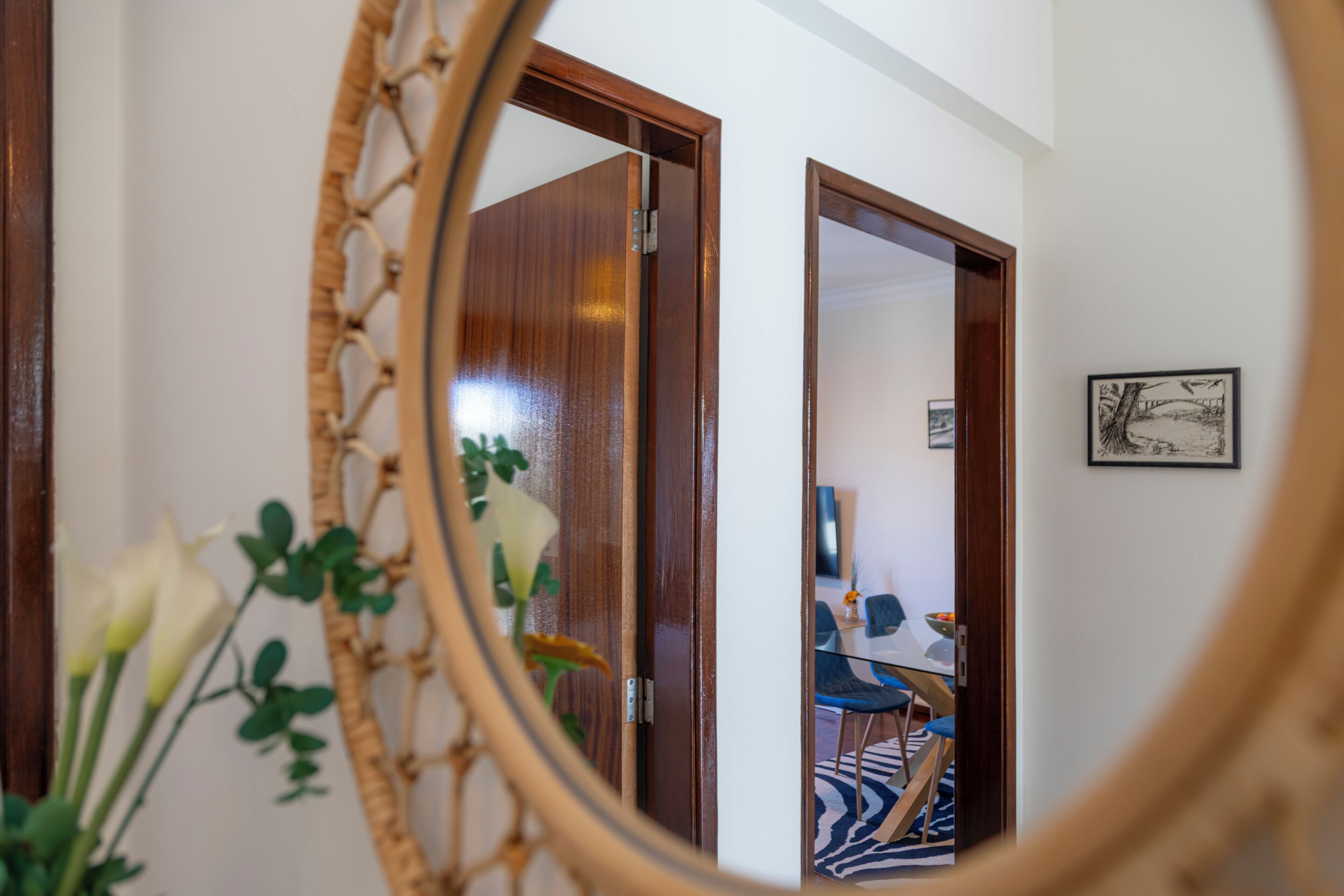 A room with a wooden door, a framed picture on the wall, and a mirror reflecting a dining area with chairs and a table.