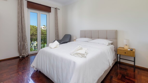 A hotel room with a large bed, a chair, a nightstand, and a view through a window. - Sea Breeze Apartment - D. Ana Beach (Lagos)
