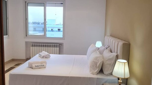 A hotel room with a bed, a nightstand, a lamp, a window, and a view of the city. - Luxurious 3-bedroom Apartment in Heart of Tunis (Tunis)