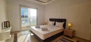 Habitación de hotel con una cama grande, dos lámparas de noche, vistas a través de un ventanal corredero y una pequeña mesa con lámpara.