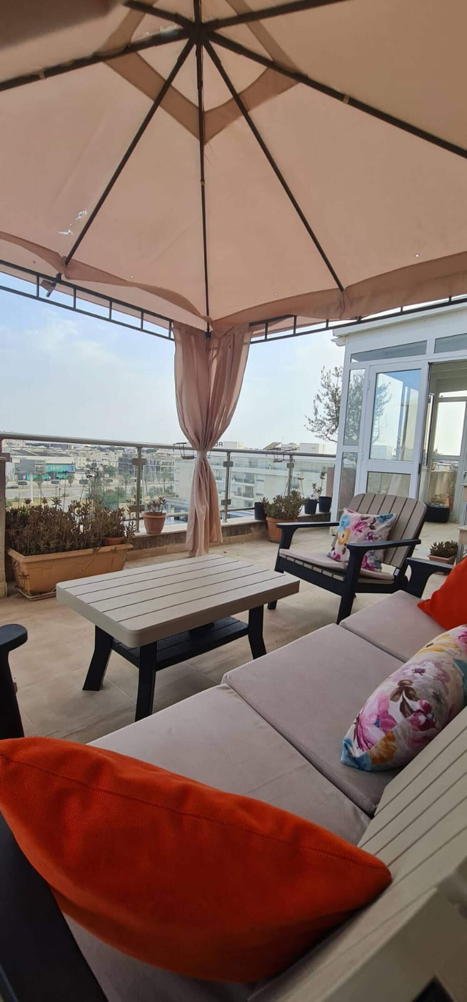 A rooftop terrace with a table and chairs, a large umbrella, and potted plants.