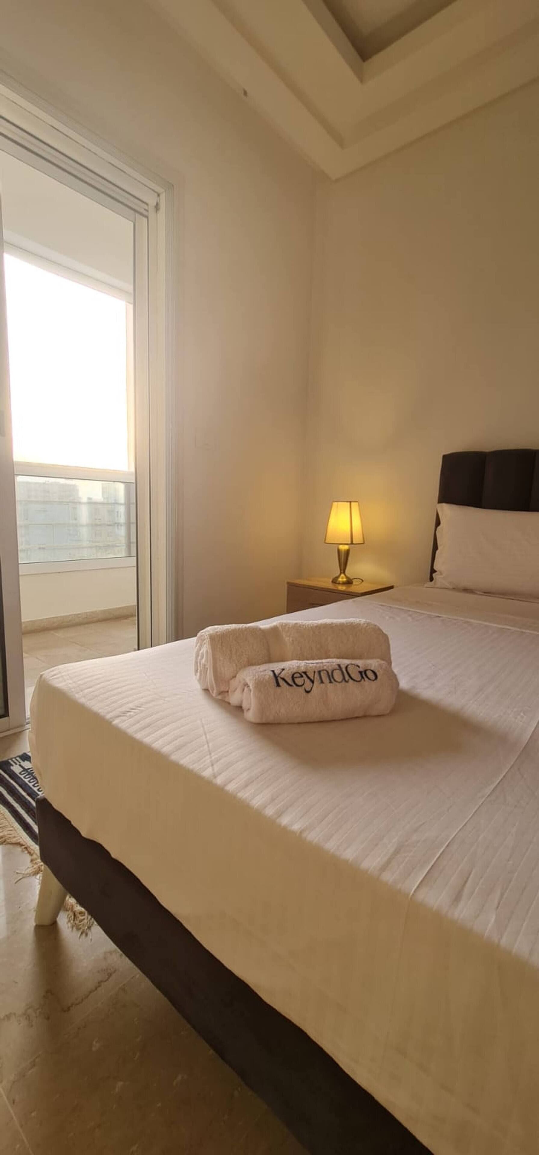 Ein Hotelzimmer mit einem Bett, einem Nachttisch mit Lampe, einem Fenster und einem Handtuch mit dem Aufdruck 'KeyndGo'.