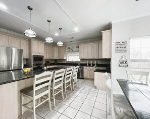 A modern kitchen with white cabinetry, a central island with bar stools, stainless steel appliances, pendant lighting, and a 'Family Rules' sign on the wall. - Resort-Style 
Backyard Retreat 
Pool-Spa & Firepit (Garden Ridge)