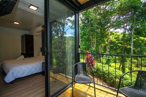 Ein Schlafzimmer mit Bett und Nachttisch, ein Balkon mit Stuhl und Blick auf einen Wald.