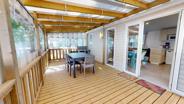 A covered outdoor deck with a wooden table and chairs, a sliding glass door leading to a kitchen, and a view of the outdoors. - Sun & Pool: 3-bedroom mobile home, patio & pool (Saint-Hilaire-de-Riez)