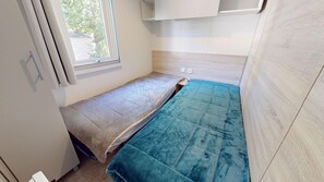 A small, narrow room with two beds, a window, and a light-colored wall. - Sun & Pool: 3-bedroom mobile home, patio & pool (Saint-Hilaire-de-Riez)