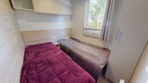 A compact room with a bed, a small table, and a window. - Sun & Pool: 3-bedroom mobile home, patio & pool (Saint-Hilaire-de-Riez)