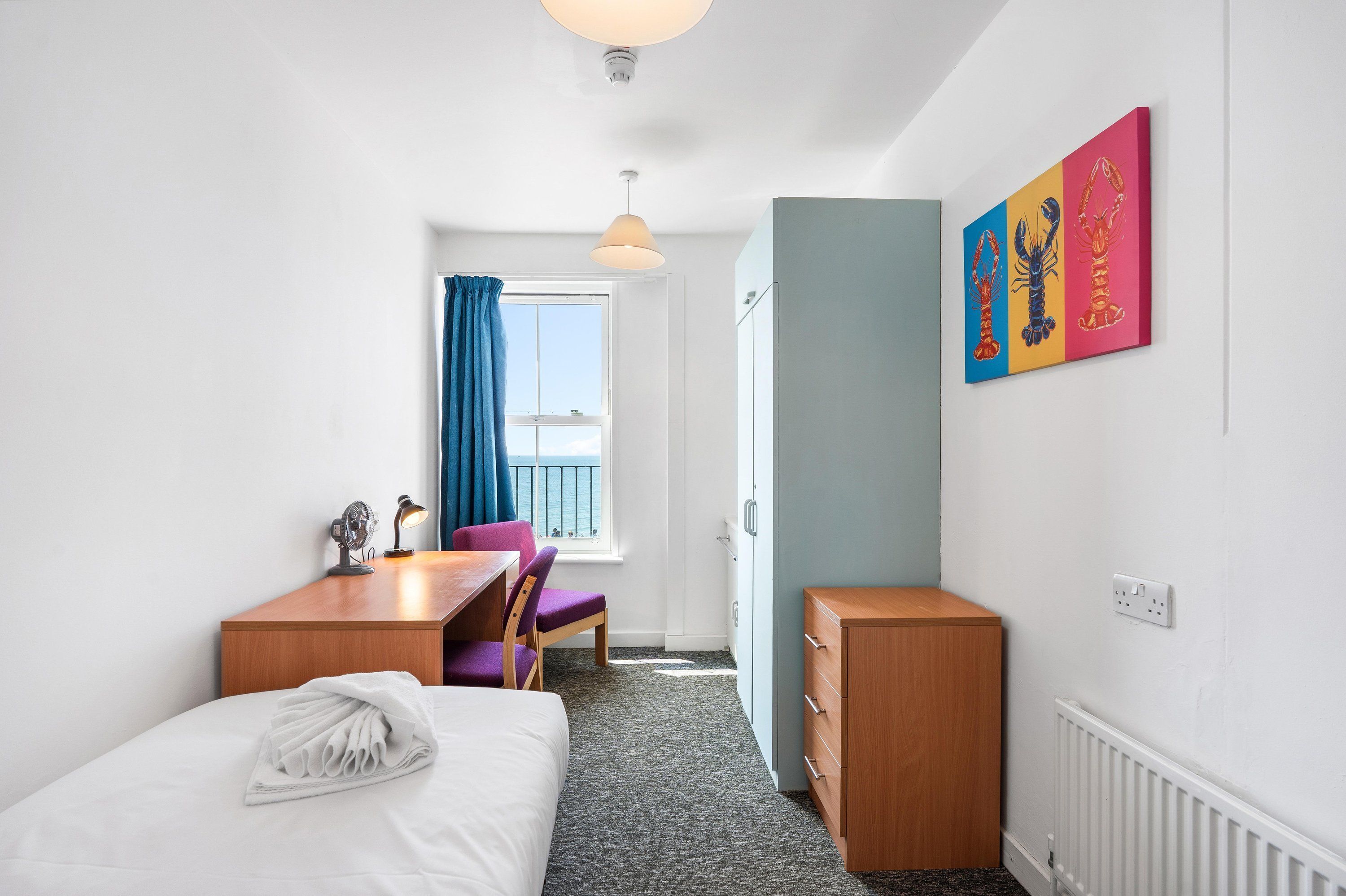 A hotel room with a bed, a desk, a chair, a radiator, a window with curtains, and artwork on the wall.