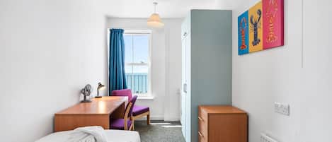A hotel room with a bed, a desk, a chair, a radiator, a window with curtains, and artwork on the wall.