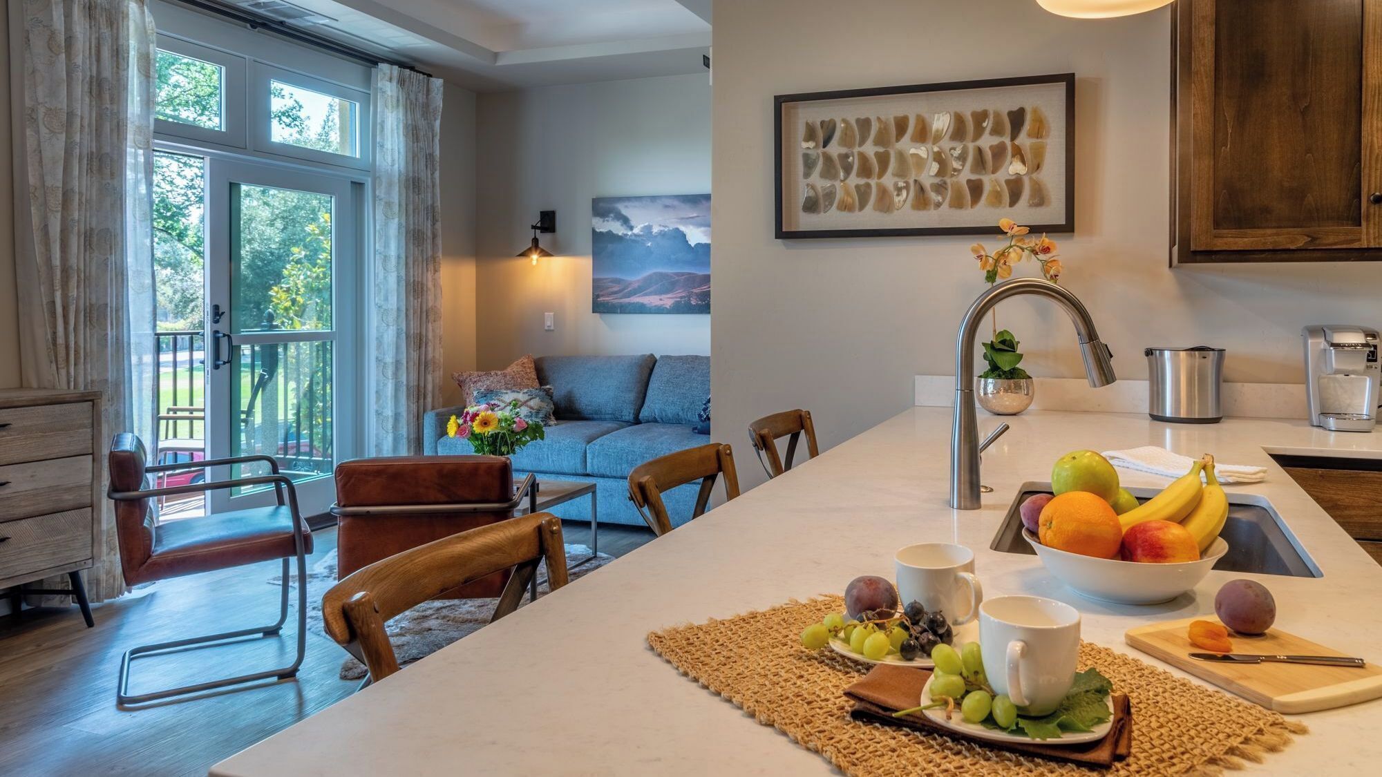 Une cuisine moderne avec un îlot central, un coin repas avec des chaises et un salon avec un canapé et un tableau.