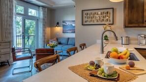 Uma cozinha moderna com ilha central, área de jantar com cadeiras e sala de estar com sofá e pintura.