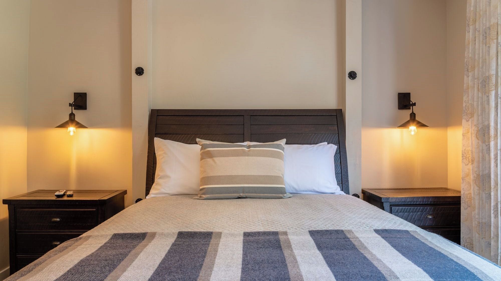 Een bed met een gestreepte deken, twee nachtkastjes met lampen, en een donker houten hoofdeinde.