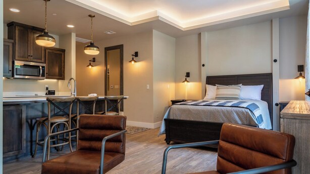 A modern hotel room with a kitchenette, a bed with a striped duvet, and two leather chairs. - Paso Park Suites by Avantstay 2 Private Balconies + Town Square Views Downtown Paso Robles (Paso Robles)