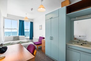Une chambre d’hôtel comprenant un lit, un bureau, une chaise, une armoire, une salle de bain avec un lavabo et un miroir, et une vue sur la mer depuis la fenêtre.