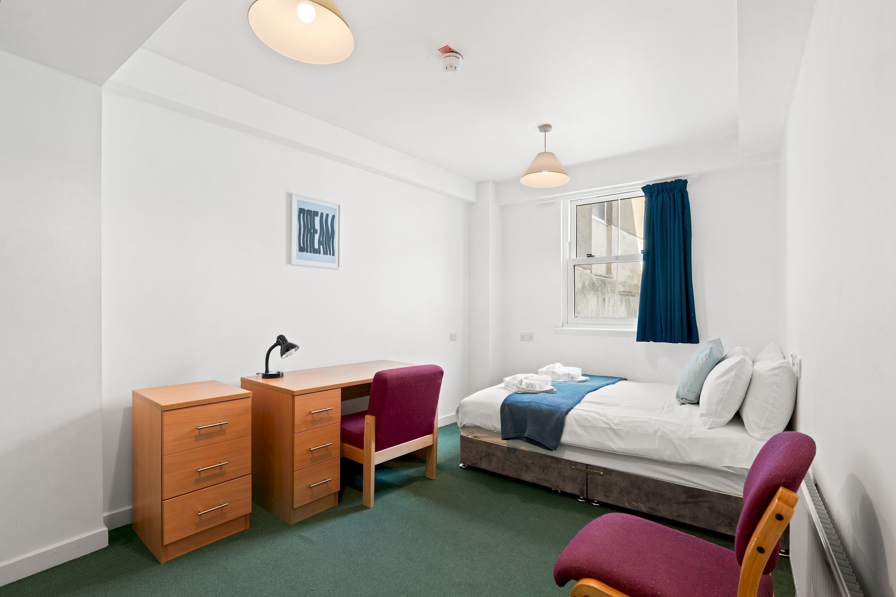 A small, well-lit room with a bed, a desk, and a chair.