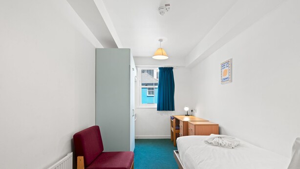 A small, well-lit room with a bed, a desk, a chair, and a window with curtains. - Kings Residence 4 | By My Getaways (Brighton)