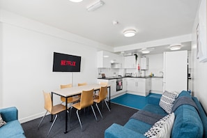 A modern living room with a sofa, a dining table with chairs, a kitchen area with cabinets and appliances, and a mounted television displaying 'NETFLIX'. - Kings Residence 4 | By My Getaways (Brighton)