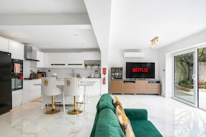 A modern kitchen with white cabinetry, a dining area with a white table and chairs, a green sofa in the living room, and a large flat-screen TV mounted on the wall. - Downs Heights | By My Getaways (Brighton)