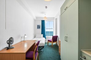 Ein Hotelzimmer mit Bett, Schreibtisch mit Ventilator, Stuhl, Kleiderschrank und Fenster mit Meerblick.