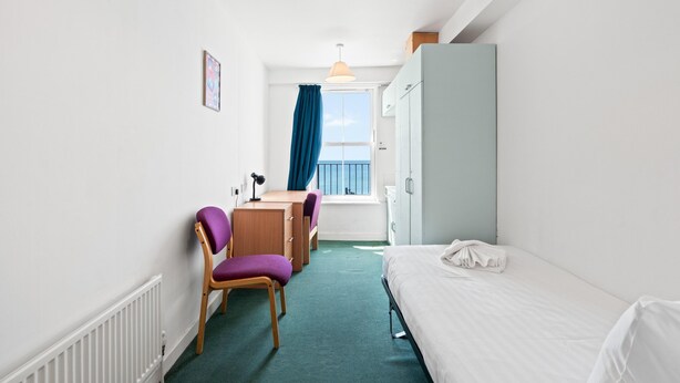 Hotellihuone, jossa on sänky, työpöytä tuolilla, ikkuna verhoilla ja näkymä merelle.