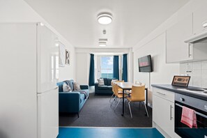 A compact hotel room with a kitchenette, a dining area with a table and chairs, a sofa, and a large window with curtains. - Kings Residence 5 | By My Getaways (Brighton)
