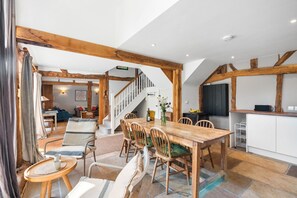 A spacious dining area with a wooden table and chairs, a staircase leading to an upper level, and a kitchen area with white cabinets. - Medleys Barn | By My Getaways (High Hurstwood)