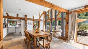 A spacious dining area with a wooden table and chairs, a kitchen in the background, and large windows with curtains. - Medleys Barn | By My Getaways (High Hurstwood)