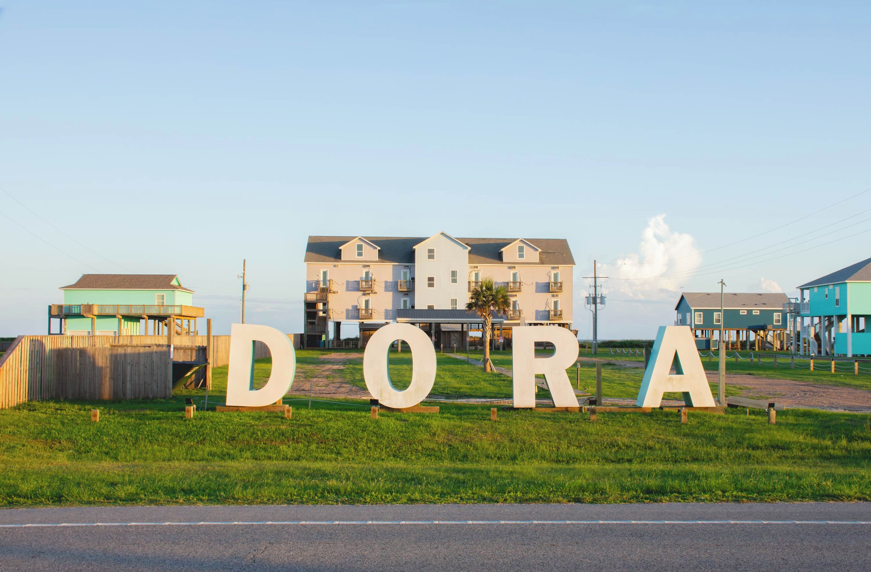 A sign with the letters 'DORA' in large letters, a grassy area, and a building in the background.