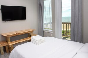 Habitación de hotel con cama, banco de madera, televisor montado en la pared y ventana con vista al mar.