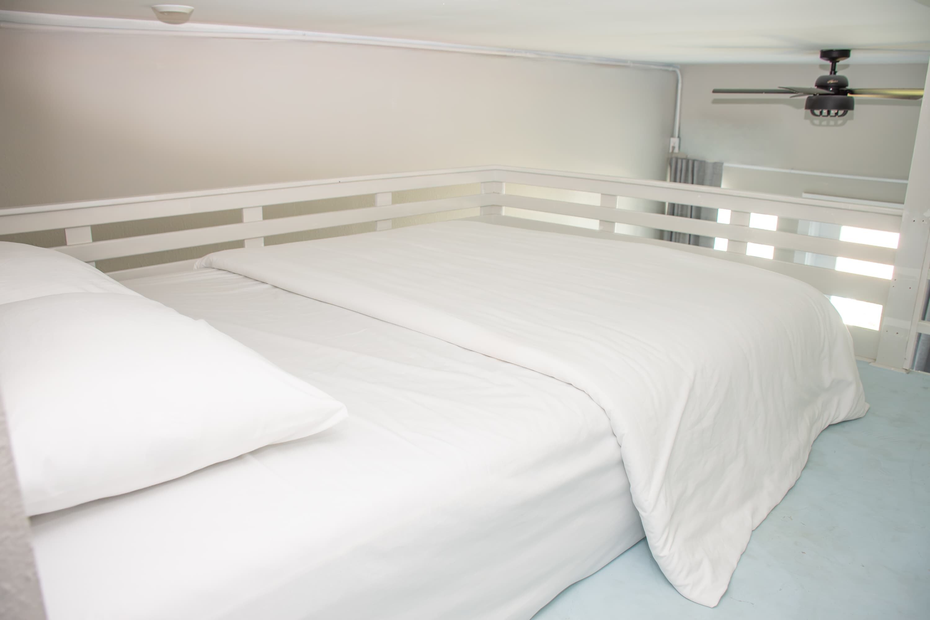 Una cama con ropa de cama blanca en una habitación con cabecera blanca y ventilador de techo.