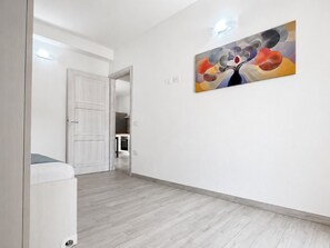 A hallway with a white door, a painting on the wall, and a view into a kitchen area. - Pegaso - Two Bedroom Apartment, Sleeps 5 (La Caletta)
