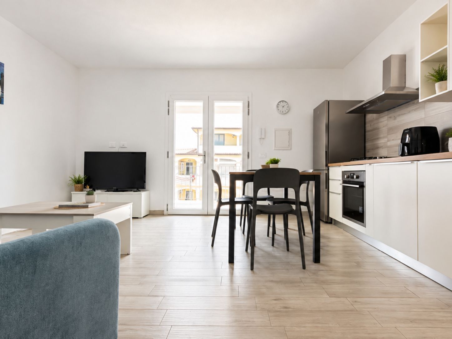 A modern kitchen with a dining area, a TV, and a view through a glass door.