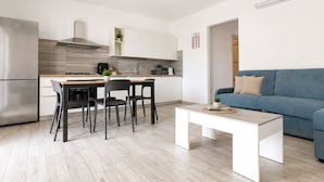 A modern kitchen with white cabinets, a dining table with chairs, and a blue sofa in the living area. - Pegaso - Two Bedroom Apartment, Sleeps 5 (La Caletta)