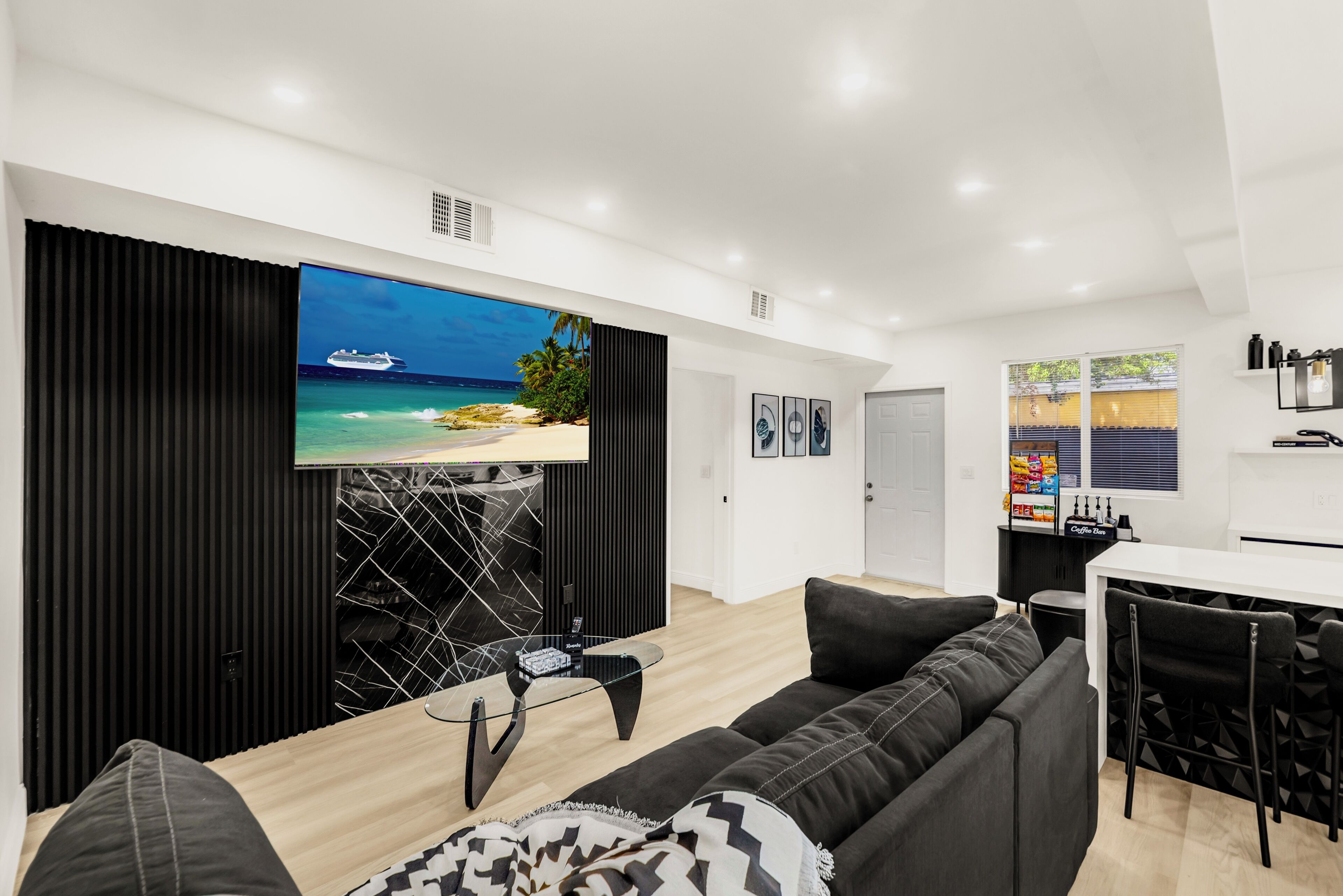 A modern living room with a large flat-screen TV mounted on a black wall, a black-framed glass coffee table, a black sofa, and a dining area with a white table and chairs.