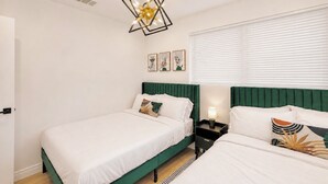 Een hotelkamer met twee bedden, een groen hoofdeinde, een moderne kroonluchter en een raam met zonwering.