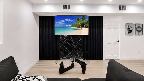 Een moderne woonkamer met een flatscreen televisie aan een zwarte muur, een glazen salontafel en een witte deur.