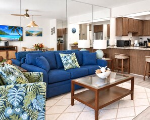 A modern living room with a blue sofa, a glass coffee table, and a kitchen area in the background. - Papakea Resort | 1-Bedroom Ocean View Villa | Car Incl. w/ 6 nights | PKD-204 by KBM (Lahaina)
