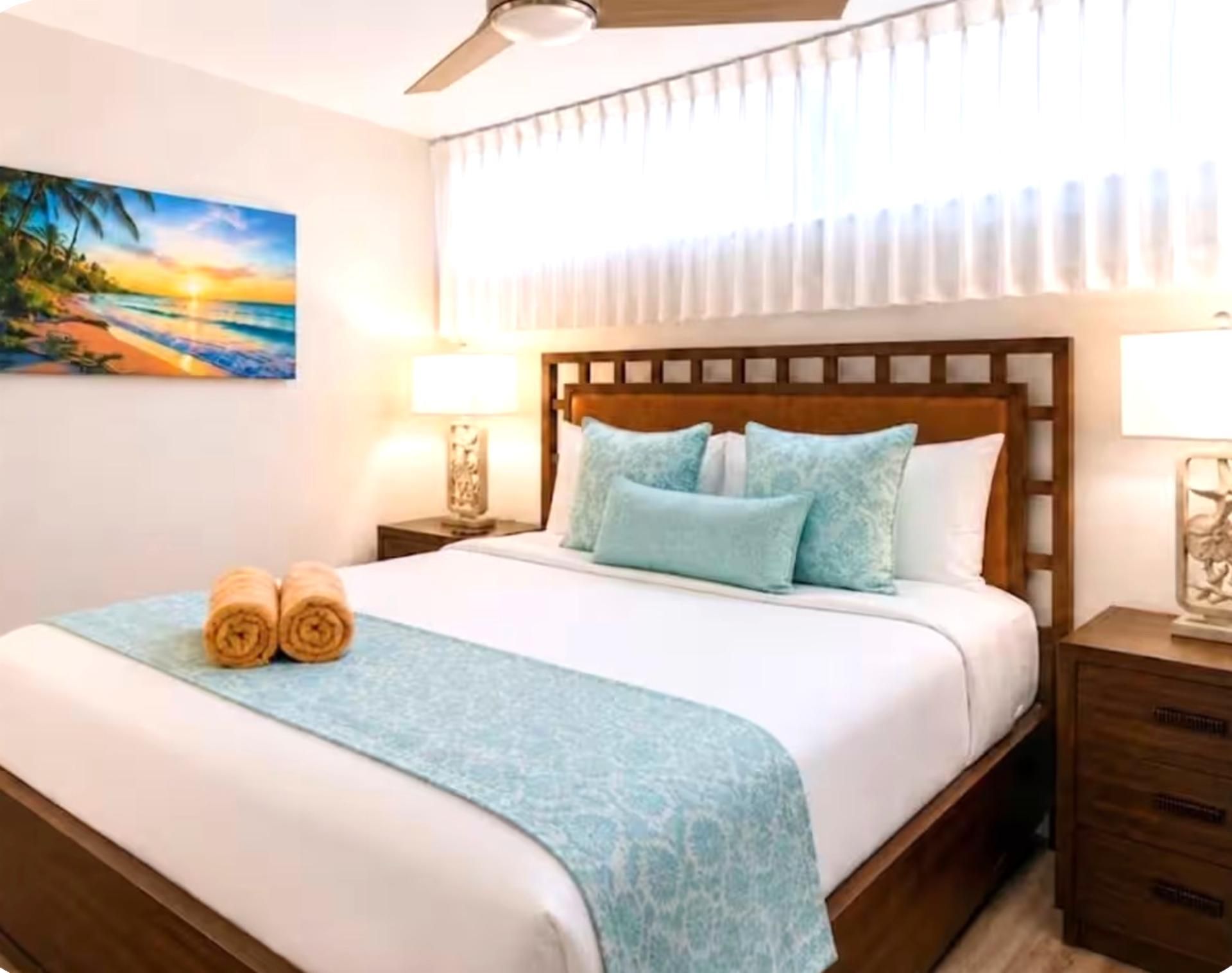 Une chambre d’hôtel avec un lit, deux lampes de chevet, un ventilateur de plafond, un tableau au mur et une fenêtre avec des rideaux.