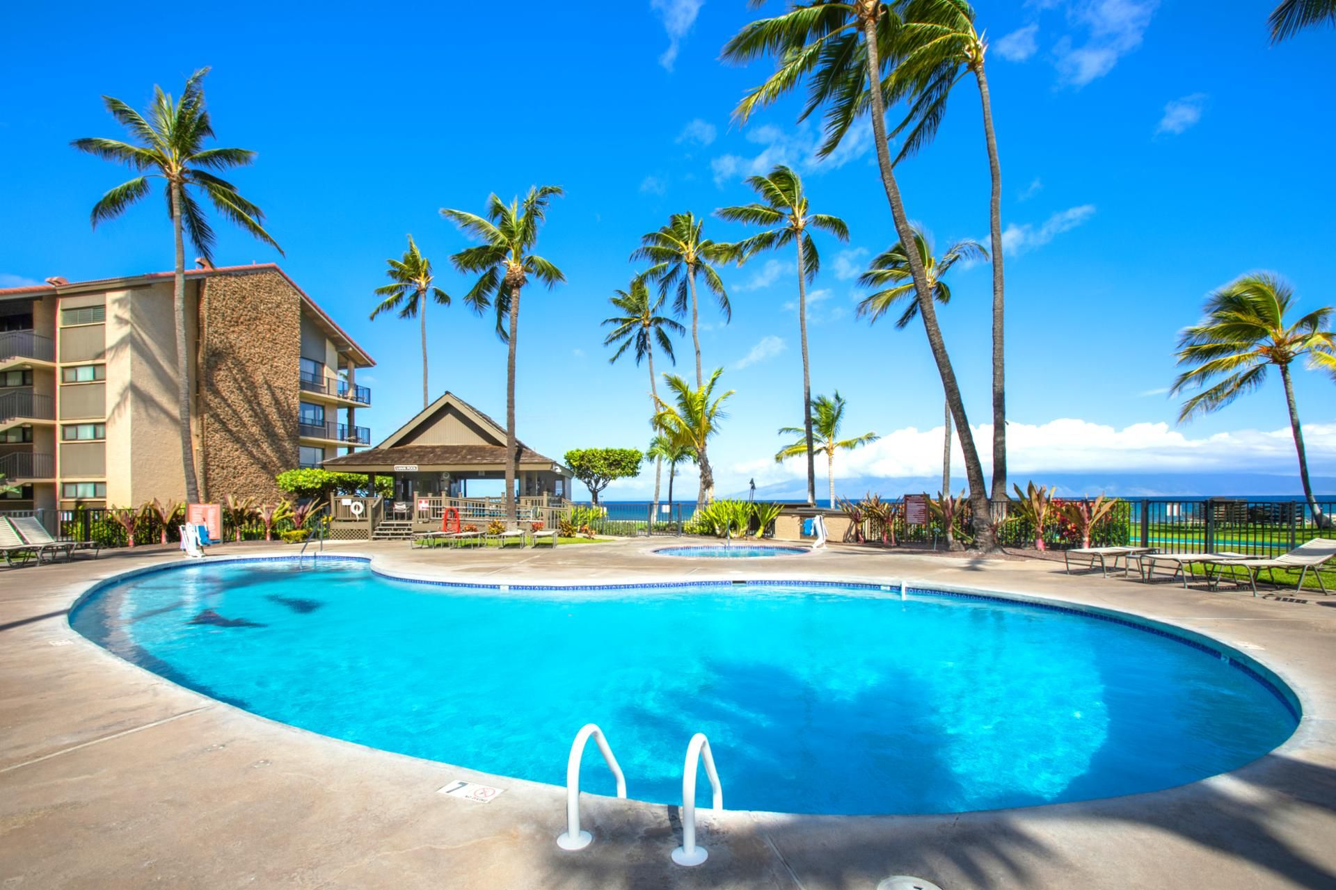 Une piscine d’un complexe hôtelier bordée de palmiers, un bâtiment avec vue sur la plage et un poste de surveillance pour les sauveteurs.