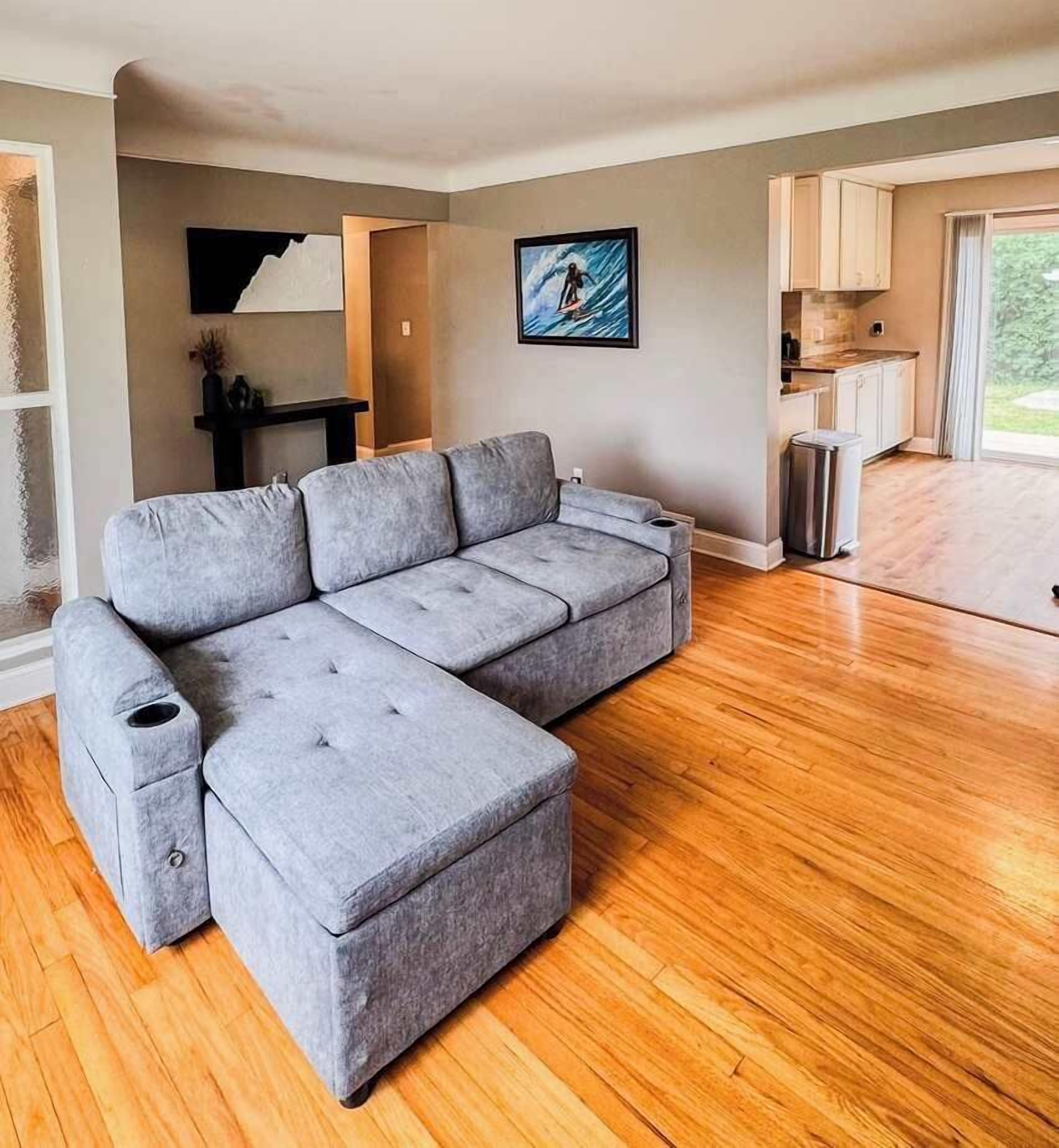 Un salon moderne avec un canapé d’angle gris, un plancher en bois et un coin cuisine visible à travers une porte ouverte.