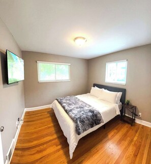 Ein Schlafzimmer mit einem Bett, zwei Fenstern, einer Deckenleuchte und einem Fernseher an der Wand.