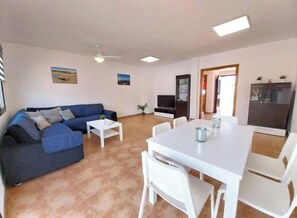 A living room with a sofa, a dining table with chairs, a TV, and a refrigerator. - Welcome, EstrellaMarina, to Corralejo, Fuerteventura! (Geafond)