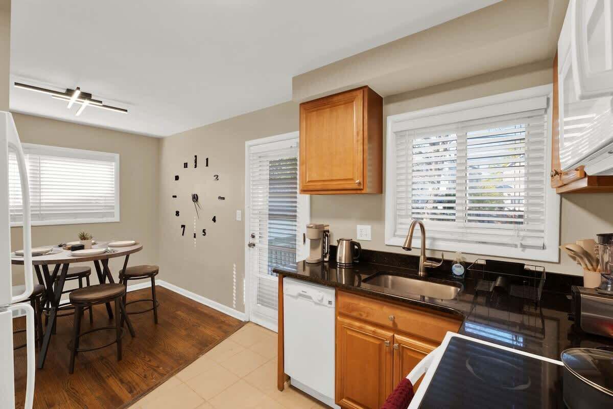 A compact kitchen with a dining area, wooden cabinets, a dishwasher, and a window with blinds.