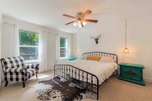 A bedroom with a bed, a chair, a ceiling fan, a mounted deer head, and a nightstand with a lamp.