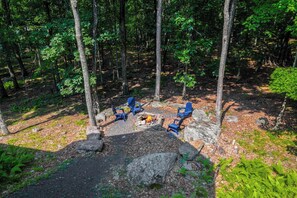 A forest campsite with a fire pit and four blue chairs arranged around it.
